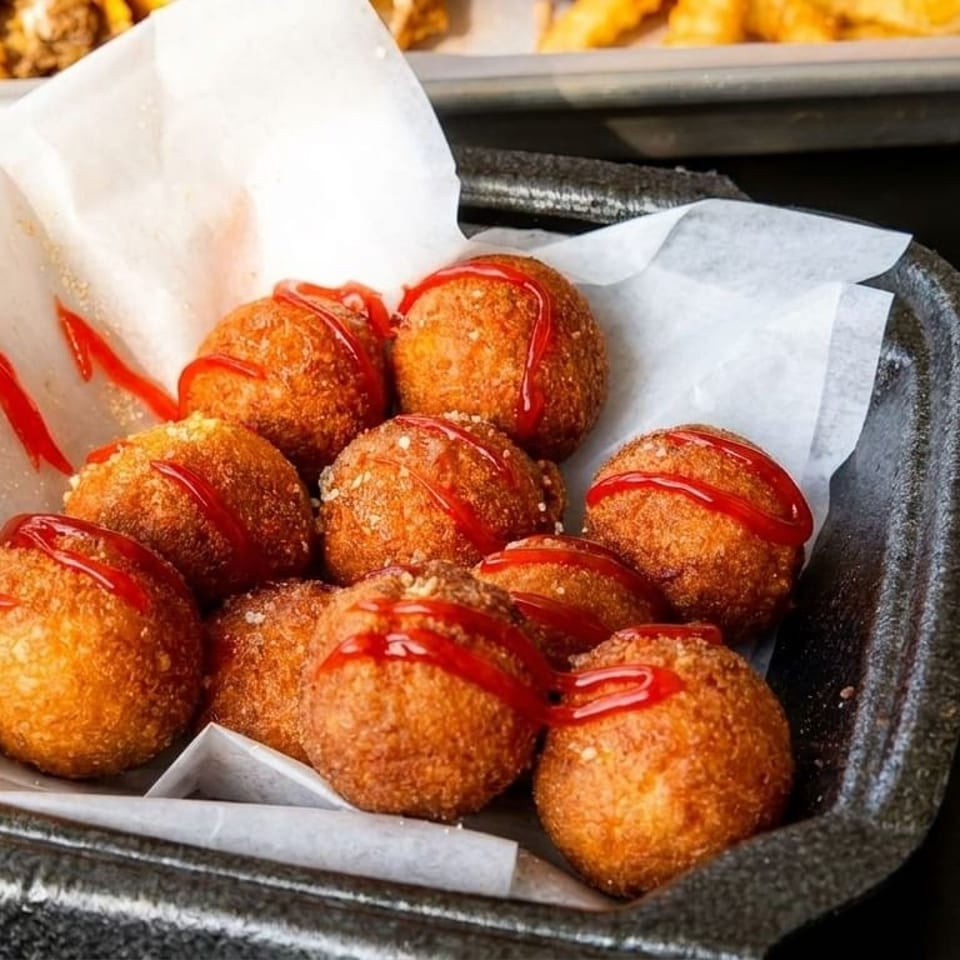Golden-brown fried hushpuppies drizzled with ketchup in a black container lined with white paper.