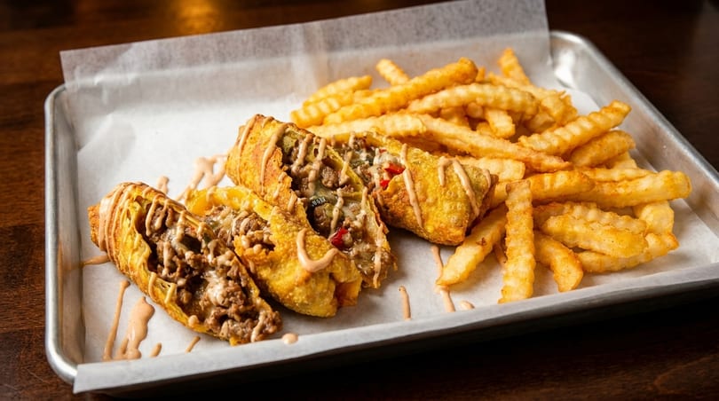 Cheesesteak egg rolls drizzled with sauce, served with crinkle-cut fries on a metal tray.