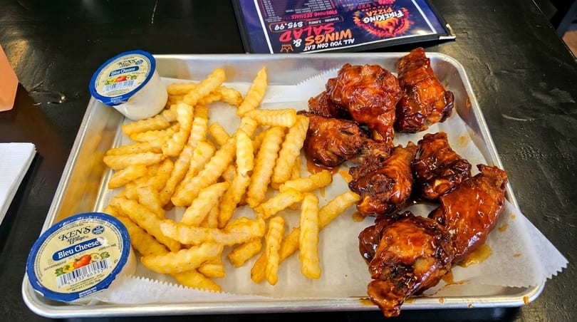 A metal tray holds crinkle-cut fries, saucy chicken wings, and two cups of Ken's Bleu Cheese dressing.