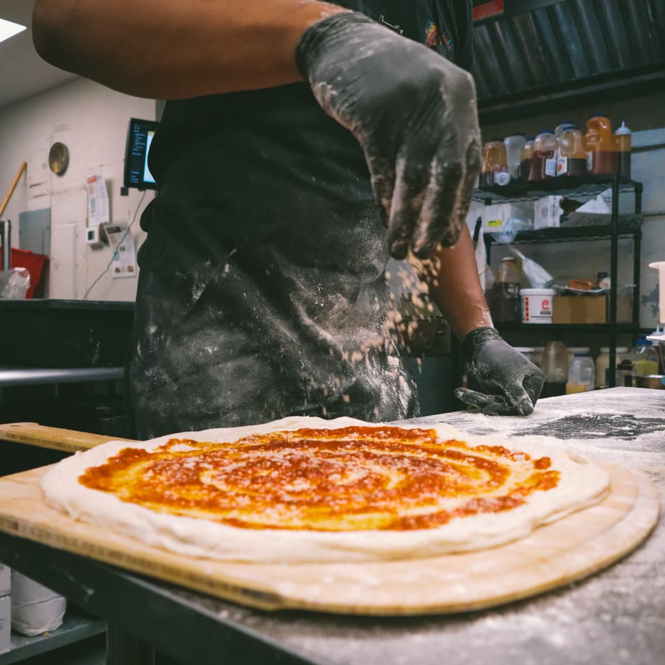 A chef in black gloves sprinkles cheese onto a pizza with red sauce on a wooden peel.