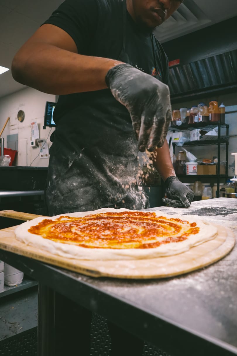 A chef in black gloves sprinkles cheese onto a pizza with red sauce on a wooden peel.