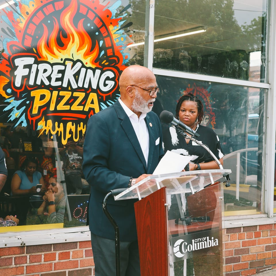 A man speaks at a podium outside Fireking Pizza, with a woman standing behind him.