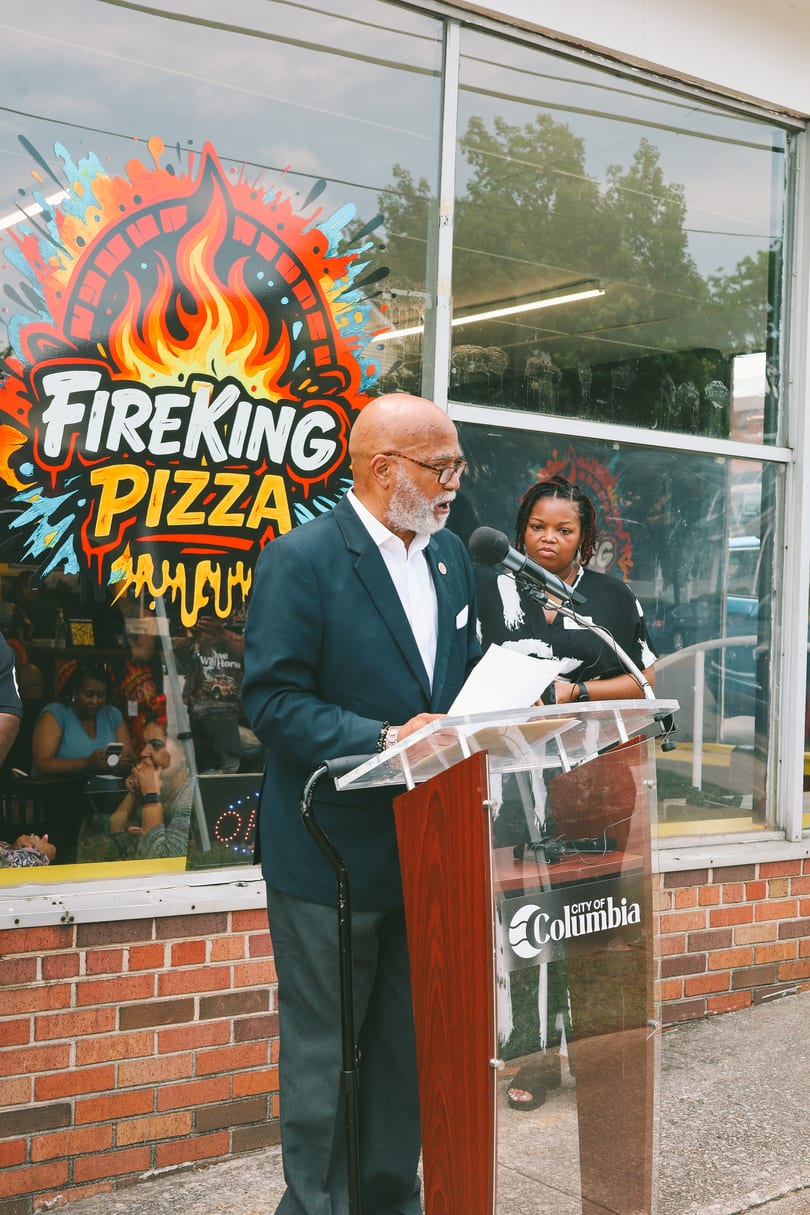 A man speaks at a podium outside Fireking Pizza, with a woman standing behind him.