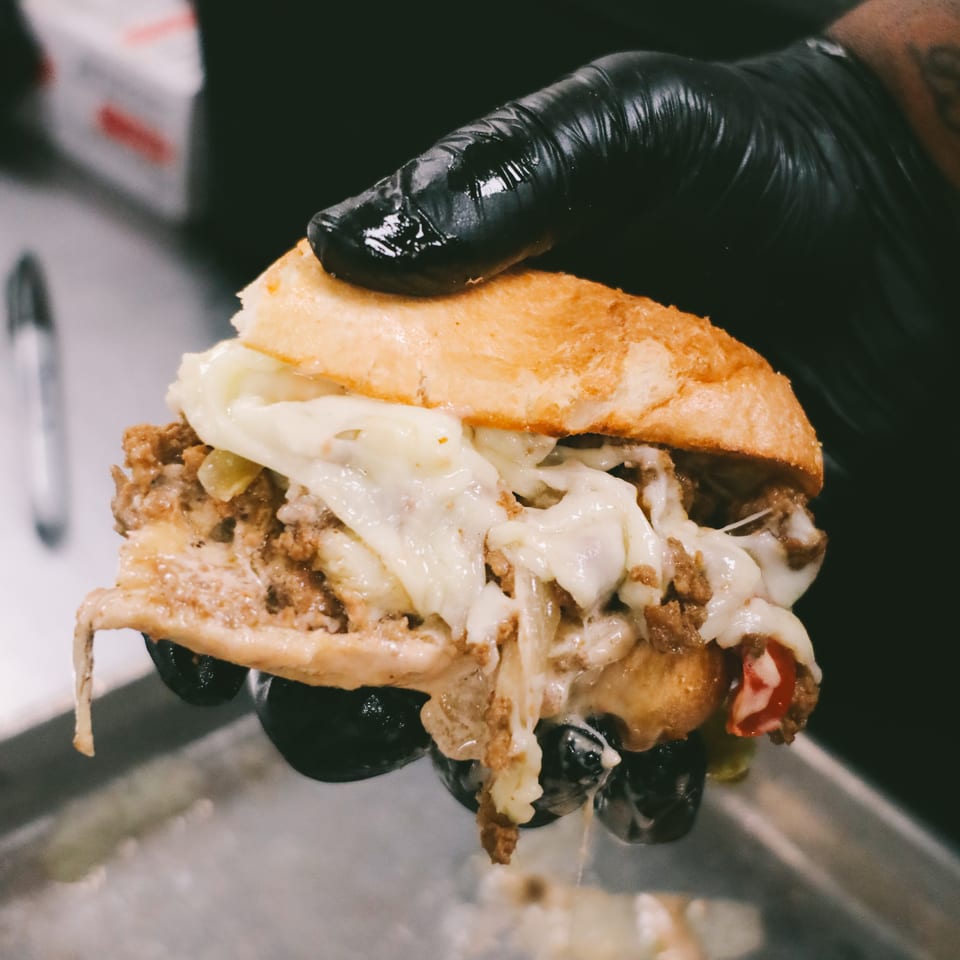 A gloved hand holds a cheesy steak and pepper sandwich over a metal tray.