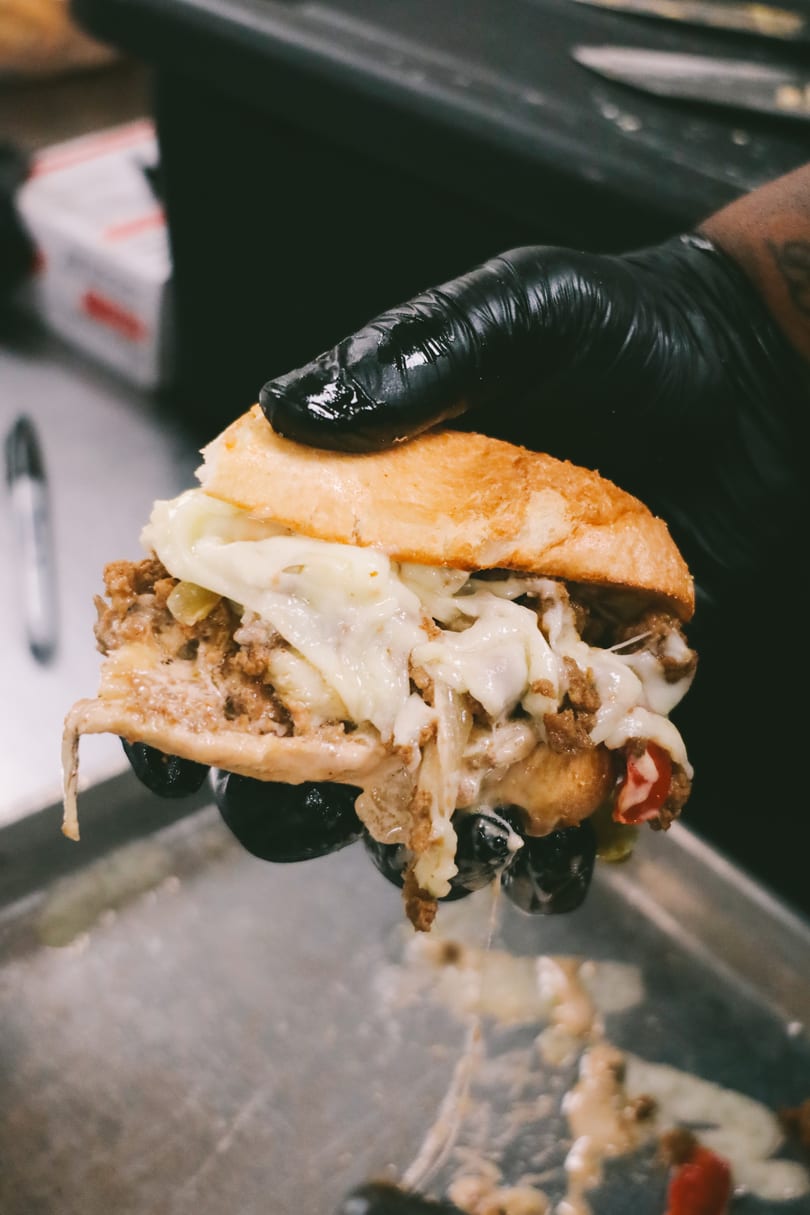 A gloved hand holds a cheesy steak and pepper sandwich over a metal tray.