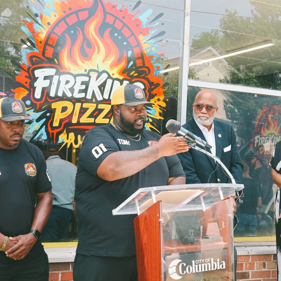 A man speaks at a podium with a microphone, flanked by two other men, in front of a FireKing Pizza sign.
