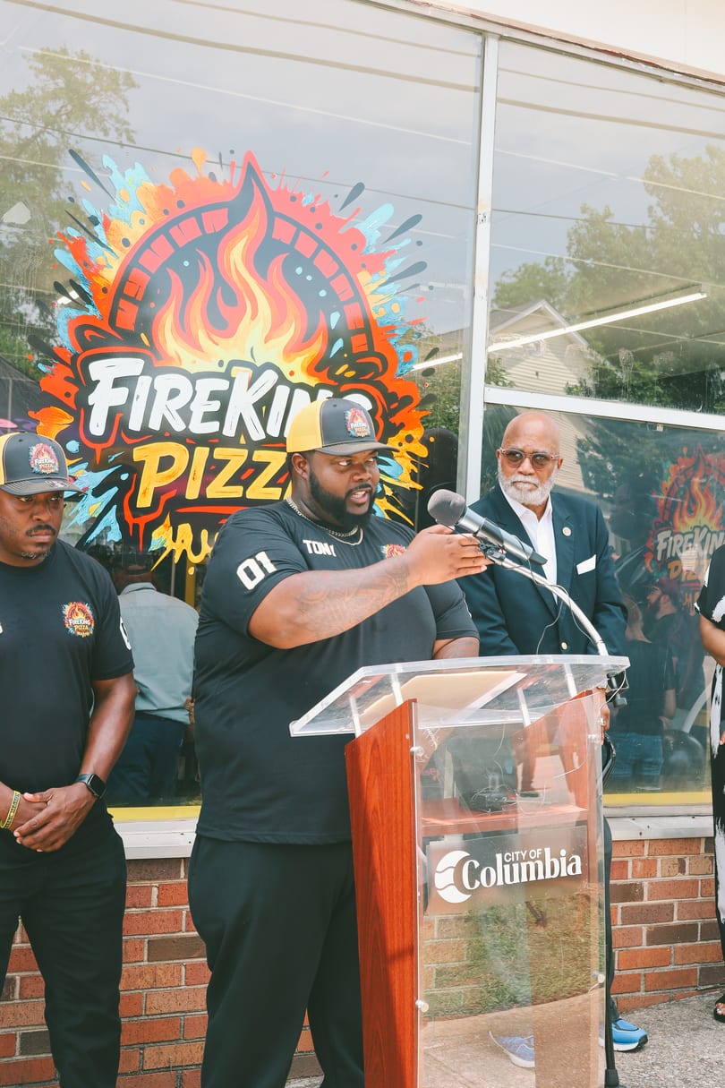 A man speaks at a podium with a microphone, flanked by two other men, in front of a FireKing Pizza sign.