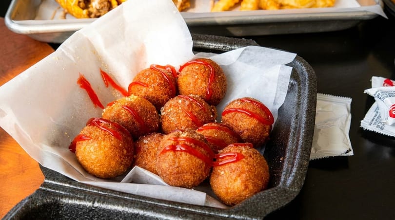 Golden-brown fried hush puppies drizzled with ketchup, served in a black container with white paper.