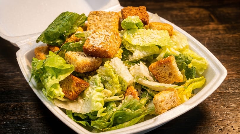 Caesar salad with romaine lettuce, croutons, and grated cheese in a white takeout container.