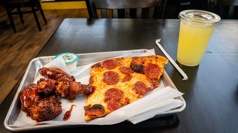 A metal tray with a slice of pepperoni pizza, BBQ chicken wings, ranch, and a lemonade.